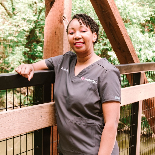 A veterinarian in gray medical scrubs leans against a wooden railing on a bridge, smiling with the serene greenery in the background.